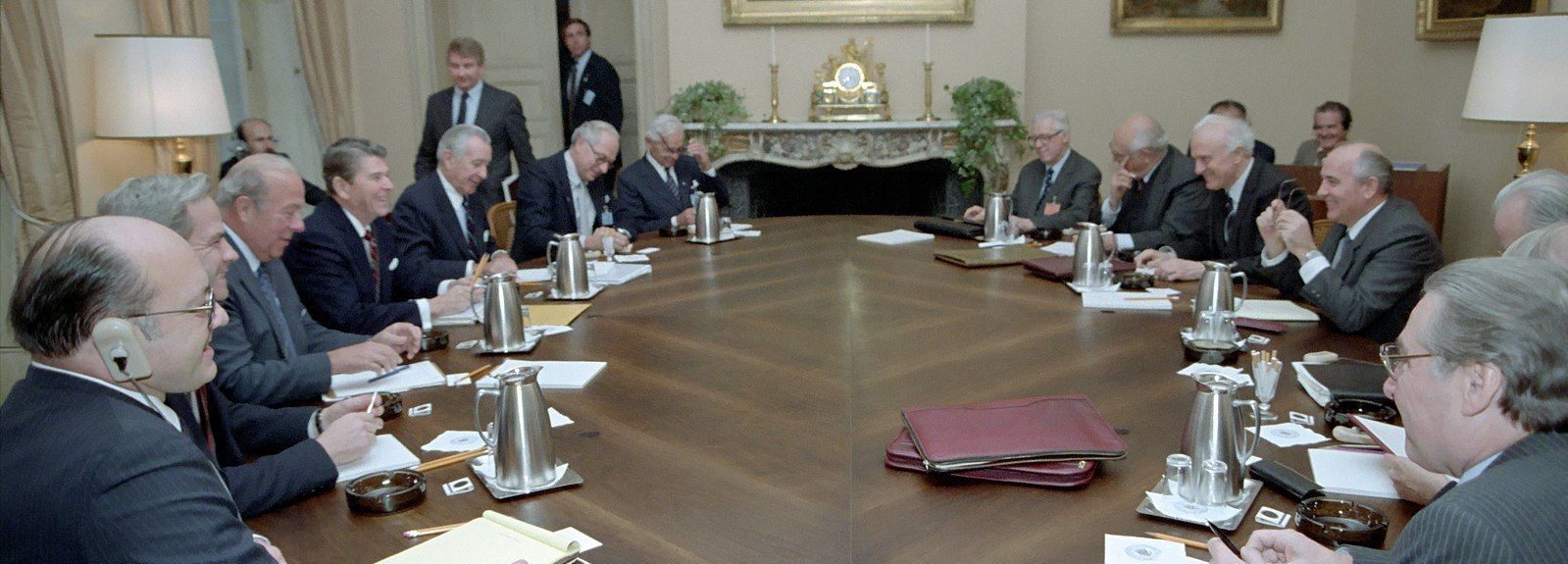 Fifteenwhite guys in ties and jackets sitting around a large oval table, with coffeepots and manila folders. Reagan and Gorbachev sit on opposite sides, both flanked by advisers.