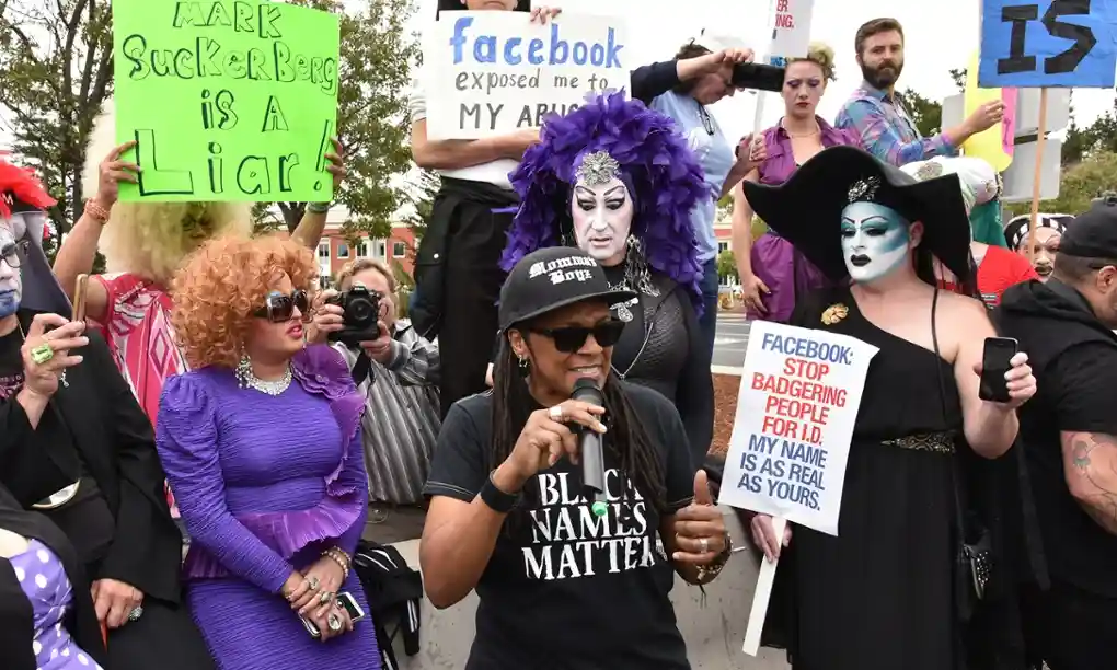 A Black person, speaking into a mic, wearing a T-shirt saying "Black names matter" and holding sign saying "Facebook: stop badering people for ID. My name is as real as yours". In the background, drag queens dressed as nuns, and other protestors, some also holding signs
