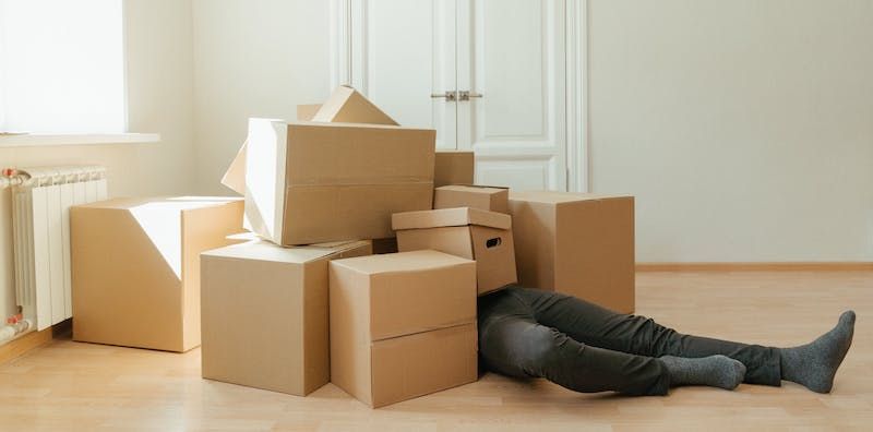Brown cardboard moving boxes on the floor, with a person beneath them whose legs and are sticking out